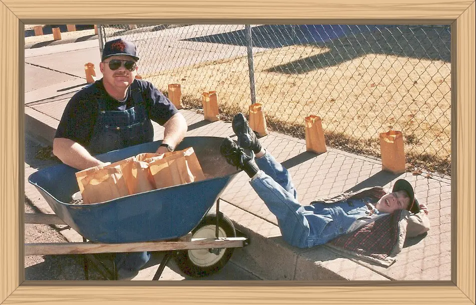 Pete and Hayes filling the paper bags with sand and adding the candles. Hayes is laying on the sidewalk with his feet resting on the wheelbarrel.