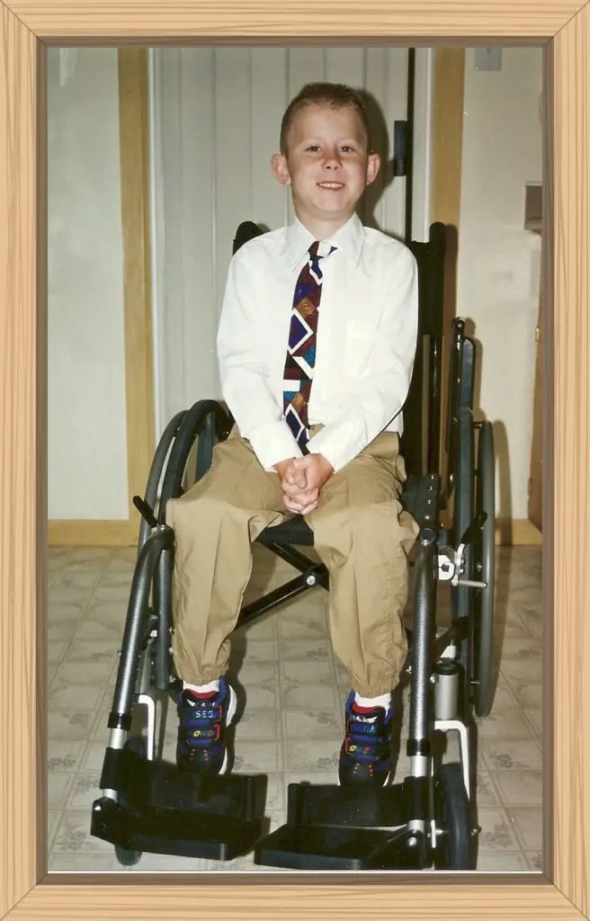 Hayes with a white shirt and tie in dress pants posing in his first manual wheelchair after he could not stand on his own any longer.