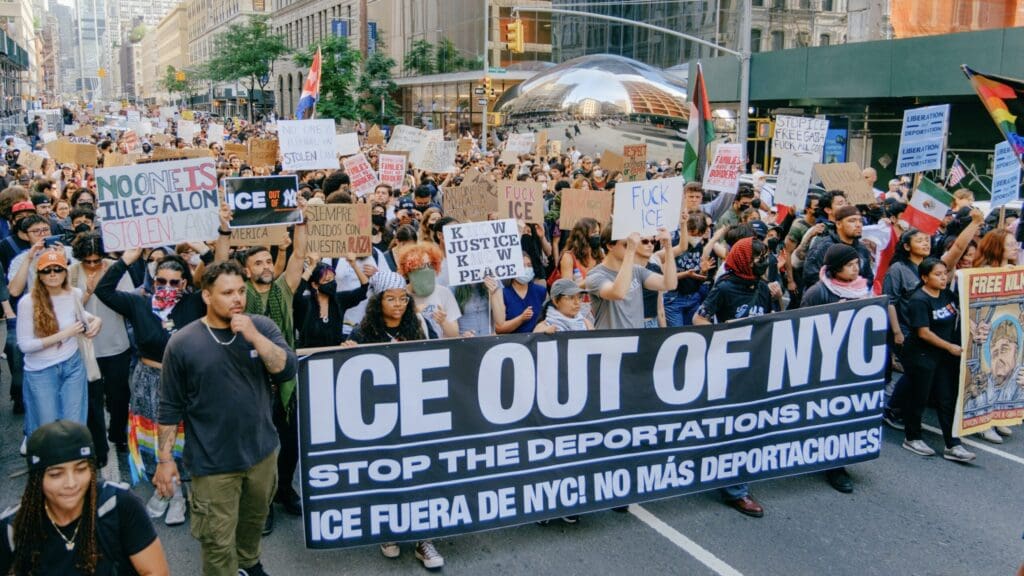 Mass protest of people in New York City carrying a large banner that says, ICE OUT OF NYC Stop The Deportations Now!